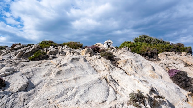 Een oud en klein heiligdom gelegen op rotsen nabij de Egeïsche Zeekust, struiken rondom, bewolkte hemel, Griekenland