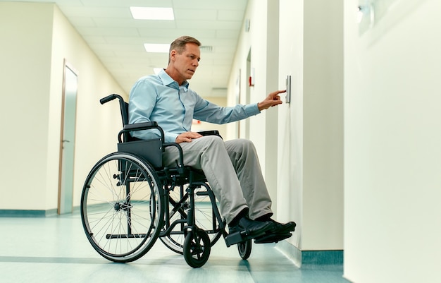 Een oudere gehandicapte man in een rolstoel drukt op de belknop voor de lift in een moderne kliniek.