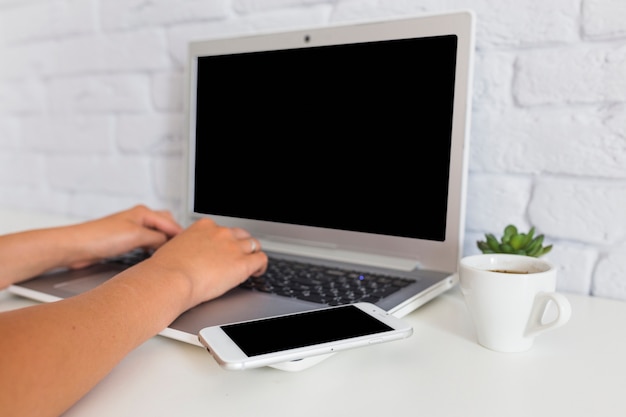 Een persoon die laptop met cellphone en koffiekop gebruikt over het bureau