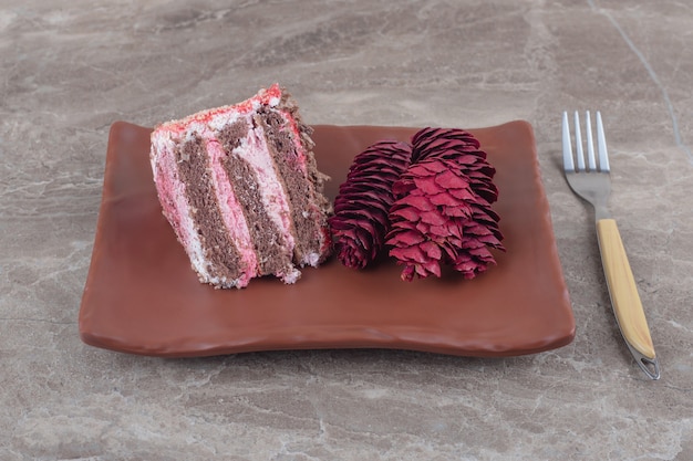 Een plakje cake en rode dennenappels op een schaal naast een vork op marmer