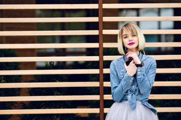 Een portret van een dromerig blond meisje met felroze lippen en naakt make-up, luisteren naar muziek op een smartphone met gestreepte houten balken achter