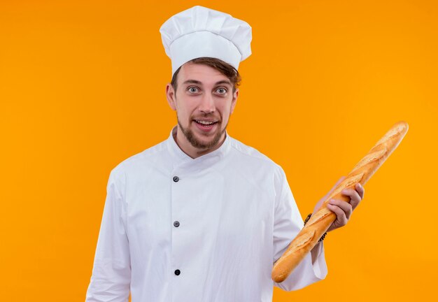 Een positieve en grappige jonge, bebaarde chef-kokmens in wit uniform die stokbrood vasthoudt terwijl hij op een oranje muur kijkt