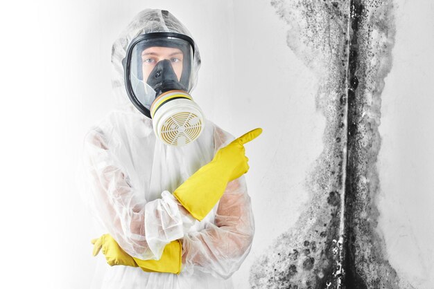 Een professionele desinfector in overall en een masker wijst met de vinger naar de zwarte schimmel aan de muur. verwijdering van de schimmel in het appartement en huis. aspergillus.