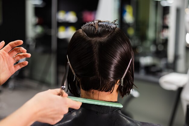 Een professionele meisjeskapper maakt het kapsel van een cliënt. Het meisje zit in een masker in de schoonheidssalon