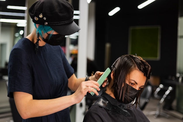 Een professionele meisjeskapper maakt het kapsel van een cliënt. Het meisje zit in een masker in de schoonheidssalon