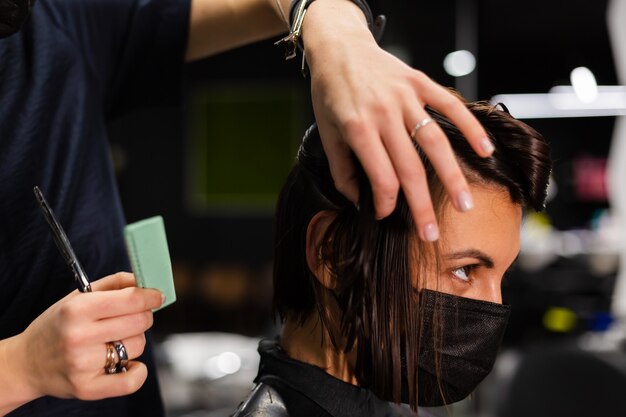 Een professionele meisjeskapper maakt het kapsel van een cliënt. Het meisje zit in een masker in de schoonheidssalon