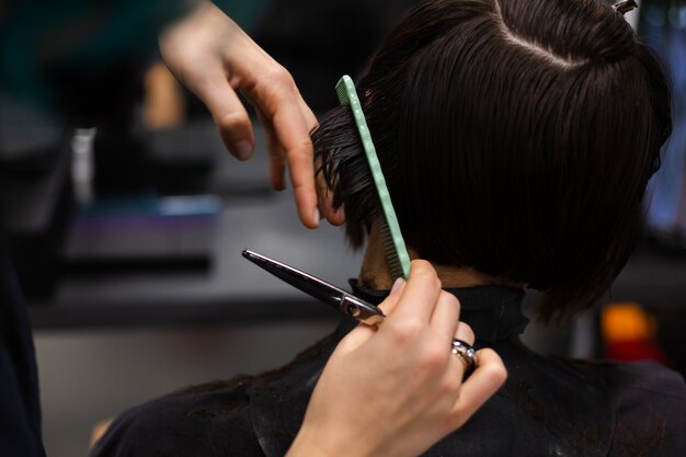 Een professionele meisjeskapper maakt het kapsel van een cliënt. Het meisje zit in een masker in de schoonheidssalon