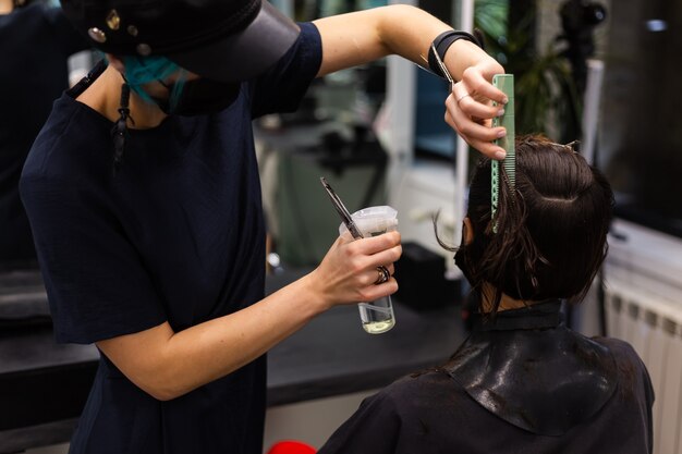 Een professionele meisjeskapper maakt het kapsel van een cliënt. Het meisje zit in een masker in de schoonheidssalon