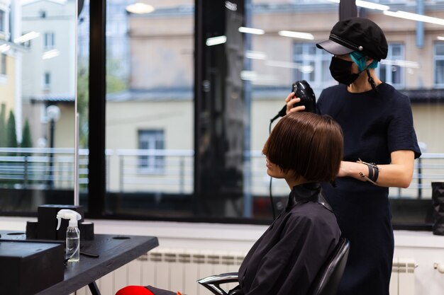 Een professionele meisjeskapper maakt het kapsel van een cliënt. Het meisje zit in een masker in de schoonheidssalon