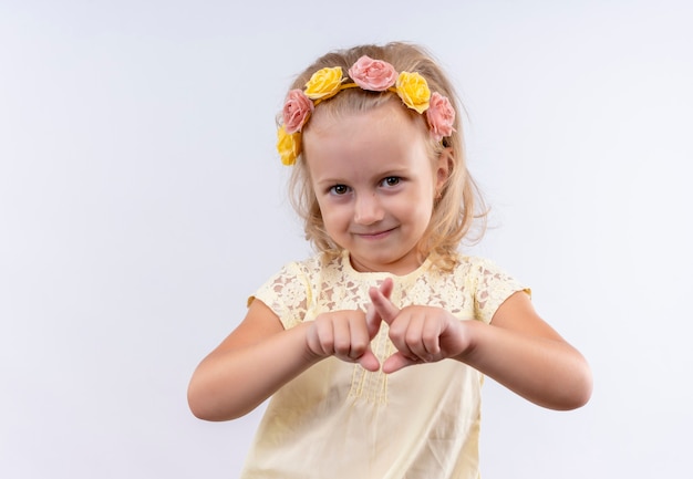 Een schattig klein meisje met een geel shirt in een bloemenhoofdband met een x-teken met vingers op een witte muur