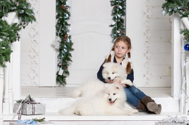Een schattig lachend kind knuffelt een witte volbloed samojeed hond puppy.blonde meisje, heeft een blij gezicht, mooie ogen, omhelst samojeed husky hond. portret vakantiehuis. winter familie dier. kindermode-stijl.