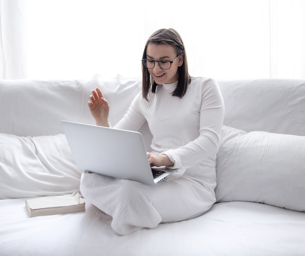 Een schattige jonge vrouw zit thuis op een witte bank in een witte jurk en werkt op een laptop.