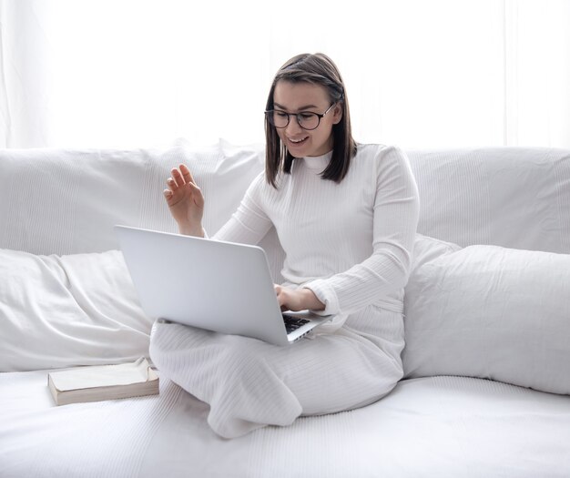 Een schattige jonge vrouw zit thuis op een witte bank in een witte jurk en werkt op een laptop.