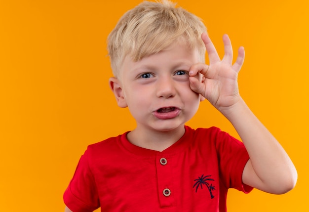 Een schattige kleine jongen met blond haar en blauwe ogen, gekleed in een rode t-shirt met ok gebaar met de hand