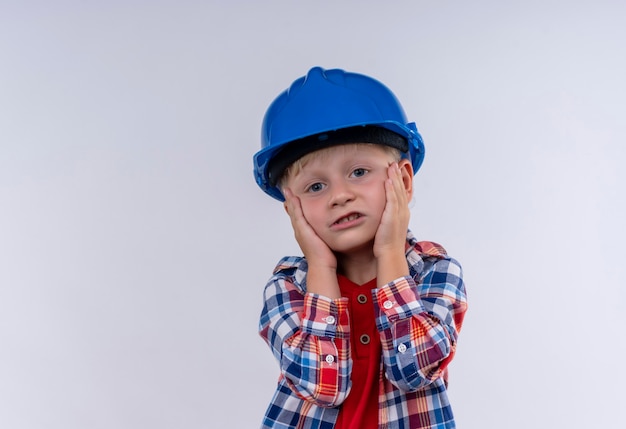 Een schattige kleine jongen met blond haar, gekleed in een geruit overhemd in een blauwe helm die handen op het gezicht op een witte muur houdt