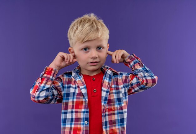 Een schattige kleine jongen met blond haar, gekleed in een geruit overhemd met de hand op de oren op een paarse muur