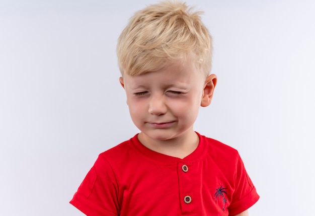 Een schattige kleine schattige jongen met blond haar, gekleed in een rood t-shirt dat zijn ogen sluit