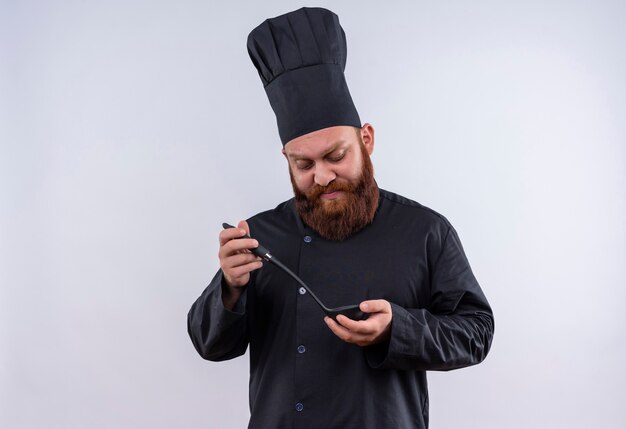 Een serieuze, bebaarde chef-kok in zwart uniform die zwarte pollepel vasthoudt en ernaar kijkt op een witte muur