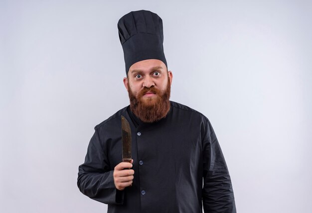 Een serieuze bebaarde chef-kok man in zwart uniform met mes terwijl hij naar de camera op een witte muur kijkt