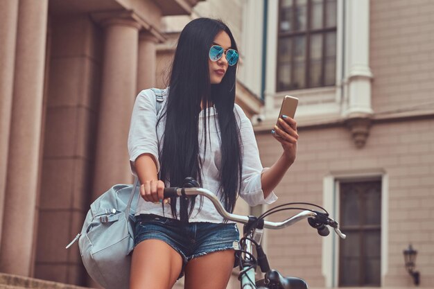 Een sexy brunette vrouw draagt blouse en denim shorts in zonnebril, leest het bericht op een smartphone, staat op trappen met een fiets in de buurt van een oud gebouw in de stad.