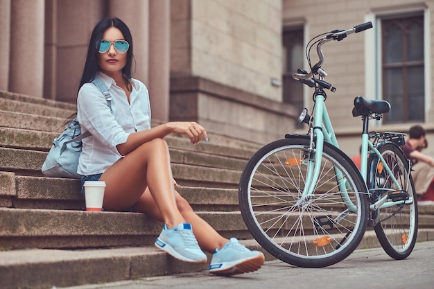 Een sexy brunette vrouw met blouse en denim shorts in zonnebrillen, ontspannen na het fietsen, zittend met een kopje koffie op trappen in een stad.