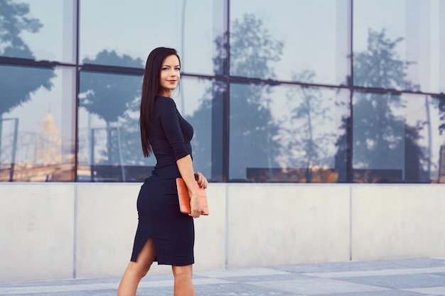 Een sexy brunette zakenvrouw gekleed in zwarte formele kleding en hoge hakken, houdt een tabletcomputer vast en gaat aan het werk, tegen een achtergrond van wolkenkrabber.