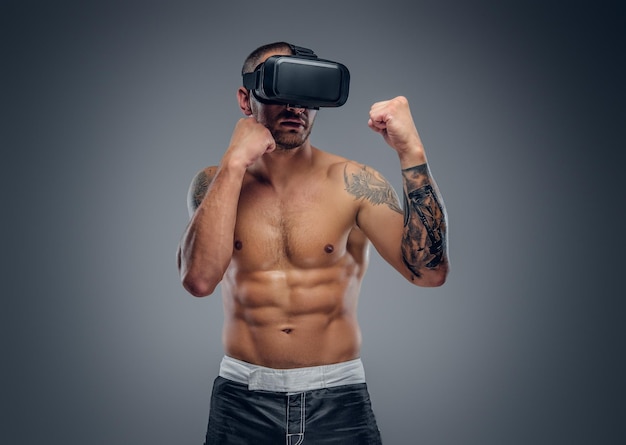 Een shirtless, getatoeëerde vechter met virtual reality-bril op zijn hoofd geïsoleerd op een grijze achtergrond.