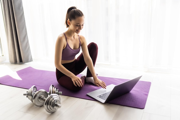 Een sportieve vrouw in sportkleding zit op de grond met een laptop thuis in de woonkamer