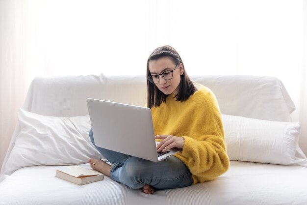 Een stijlvolle brunette meisje in een gele trui zit thuis op de bank in een lichte kamer en werkt op afstand.