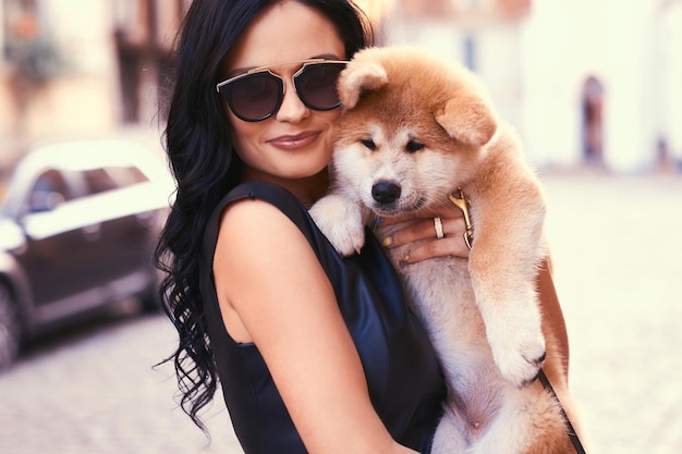 Een stijlvolle brunette vrouw in een zwarte jurk en zonnebril, staande op een straat en houdt een schattige rasechte puppy Akito vast.