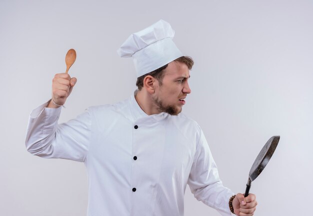 Een stressvolle jonge, bebaarde chef-kokmens in wit fornuisuniform en hoed met koekenpan en houten lepel voor aanvallen op een witte muur