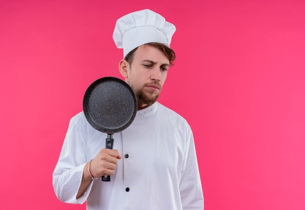 Een stressvolle jonge, bebaarde chef-kokmens in wit uniform toont koekenpan terwijl hij met droevige uitdrukking op een roze muur naar kant kijkt