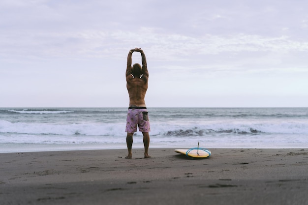 Gratis foto een surfer loopt met een plank op een zandstrand. knappe jonge man op het strand. watersport. gezonde actieve levensstijl. surfen. zomervakantie. extreme sporten.