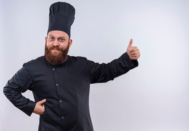 Gratis foto een tevreden bebaarde chef-kok in zwart uniform duimen opdagen terwijl hij op een witte muur kijkt