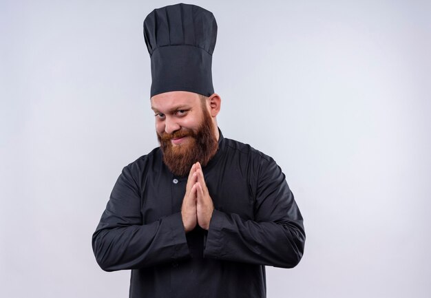 Een tevreden bebaarde chef-kok in zwart uniform hand in hand terwijl hij op een witte muur kijkt