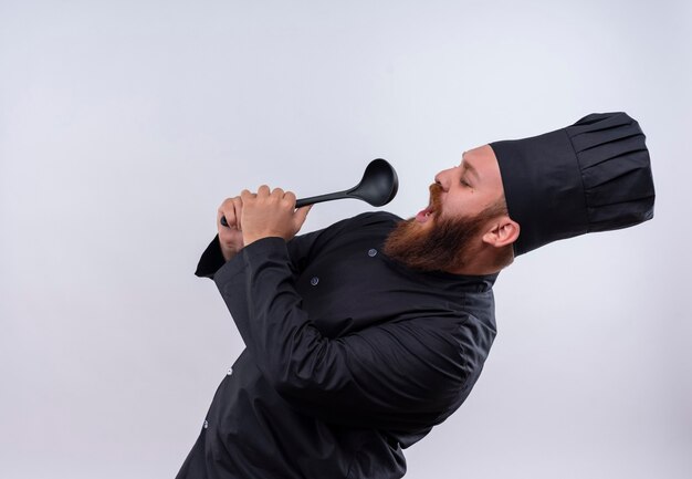 Een tevreden bebaarde chef-kok in zwart uniform zingt met pollepel en gebruikt het als een microfoon op een witte muur