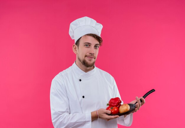 Een tevreden jonge bebaarde chef-kok in wit uniform houdt een koekenpan met verse groenten zoals ui, tomaat en paprika vast terwijl hij op een roze muur kijkt