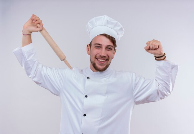 Een tevreden jonge bebaarde chef-kok man in wit fornuis uniform en hoed met deegroller met gebalde vuist terwijl hij op een witte muur kijkt