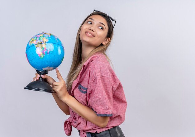 Een tevreden jonge vrouw die een rood overhemd draagt dat naar een wereldbol richt terwijl zij op een witte muur kijkt