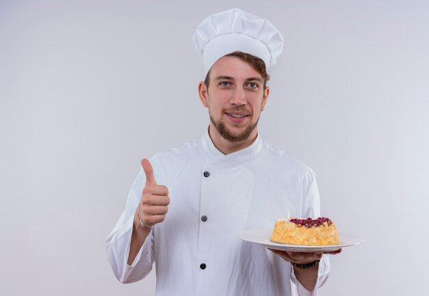 Een tevreden knappe jonge bebaarde chef-kok man in wit fornuis uniform en hoed met een bord met salade en duimen opdagen terwijl hij op een witte muur kijkt