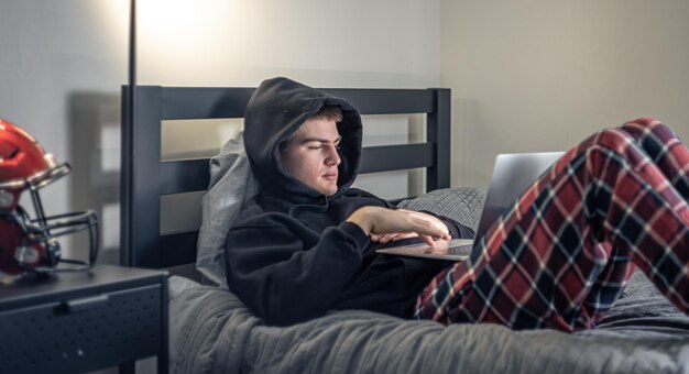 Een tienerjongen zit in een kamer op een bed en gebruikt een laptop