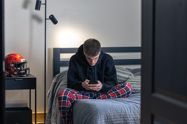 Een tienerjongen zit in een kamer op een bed en gebruikt een smartphone