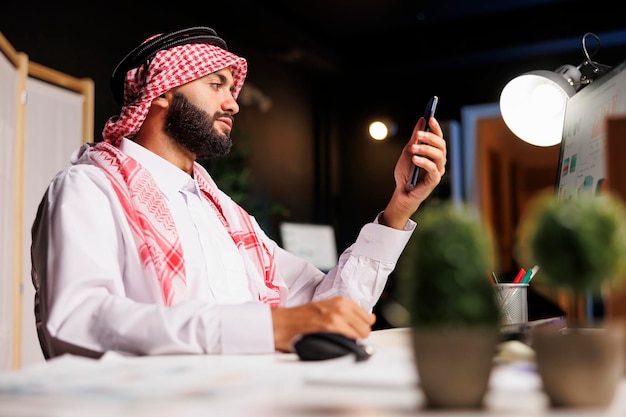 Gratis foto een traditioneel geklede zakenman uit het midden-oosten zit met zijn smartphone op een bureau in zijn kantoor. een arabische man grijpt een mobiel apparaat en gebruikt de desktopcomputer in de buurt.