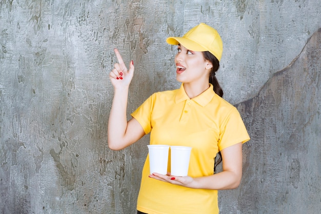 Een verkoopster in geel uniform die twee plastic bekers drank vasthoudt en naar iets wijst.