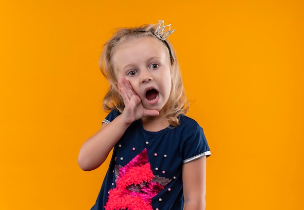 Een verrassend mooi klein meisje met een marineblauw overhemd in een kroonhoofdband die iemand belt met de handen op haar mond op een oranje muur