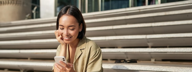 Een verticale opname van een aziatische studente zit op de trap in de stad en kijkt naar het scherm van de mobiele telefoon en