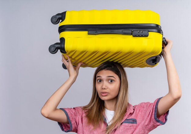 Een verwarde jonge vrouw die een rood overhemd draagt en een gele koffer op haar hoofd houdt terwijl ze op een witte muur kijkt