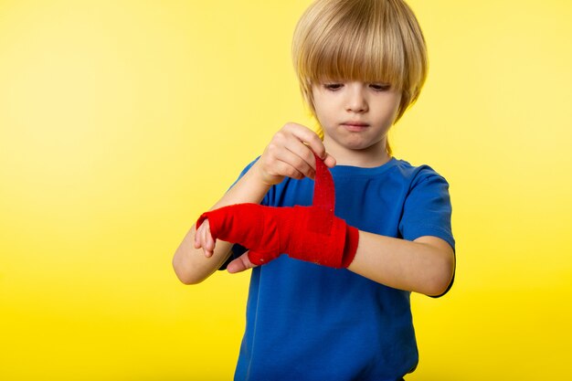 Een vooraanzicht blonde jongen schattig in blauw t-shirt en rood weefsel rond zijn hand op de gele muur