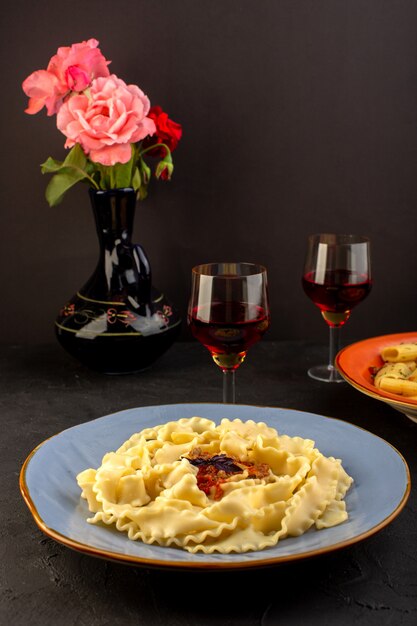 Een vooraanzicht deeg pasta gekookt lekker gezouten binnenkant ronde blauwe plaat met glazen wijn en bloemen in kruik op ontworpen tapijt en donker bureau