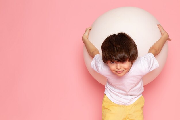 Een vooraanzicht die leuke jongen in het witte t-shirt spelen met ronde witte bal op de roze ruimte glimlachen